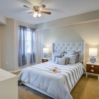 A cozy bedroom featuring a neatly made bed with striped bedding, flanked by nightstands and lamps, and softly lit by a ceiling fan light.