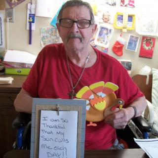 An elderly man proudly displays his Thanksgiving craft while smiling at the camera.