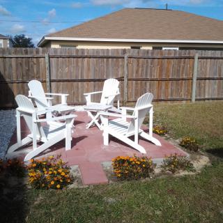 A cozy backyard patio with white chairs surrounded by vibrant flowers and a wooden fence.