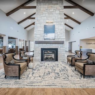 A spacious and cozy living room with a central stone fireplace, surrounded by elegant furniture and high vaulted ceilings with exposed beams.
