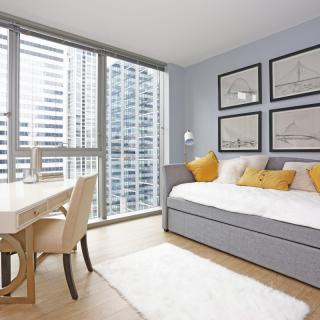 A modern apartment living space with a daybed and dining area, featuring large windows with a city view.
