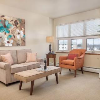 A cozy living room with a sofa, armchair, and coffee table, featuring a colorful painting and ample natural light.