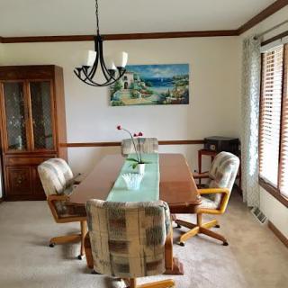 A cozy dining room with a wooden table, cushioned chairs, and a chandelier, complemented by natural light from a window.