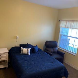 A cozy bedroom with a bed covered in blue bedding, an armchair by the window, and a nightstand beside the bed.