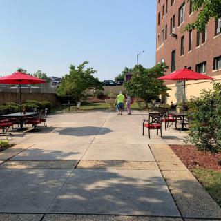 A sunny outdoor patio with tables, chairs, and red umbrellas next to a building.