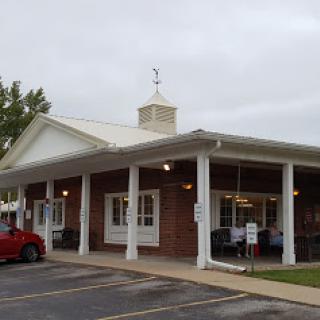 A view of a brick building with a porch and parking area, set against an overcast sky.