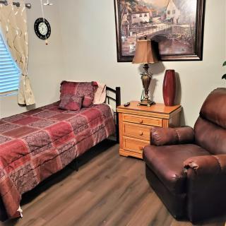 A cozy bedroom scene featuring a single bed with red plaid bedding, a dark brown armchair, and a wooden nightstand topped with a lamp. A landscape painting hangs above the nightstand, and a clock adorns the wall beside the window.