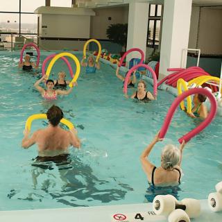 A group of people participating in a water aerobics class using colorful foam noodles in an indoor swimming pool.