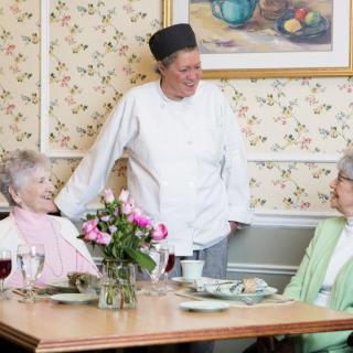 A chef engages in conversation with two elderly women seated at a dining table.