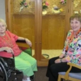 Two senior women sitting together, one in a wheelchair, indoors with decorative flowers in the background.