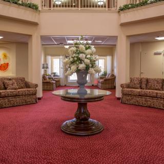 A spacious lobby area featuring a central table with a floral arrangement, surrounded by comfortable sofas and carpeted flooring.