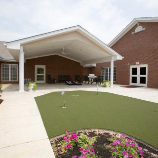 A covered patio area with a small putting green and landscaped garden in front of a brick building.