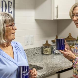 Two elderly women enjoying a conversation in the kitchen while holding mugs.