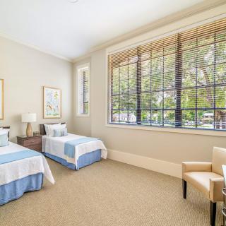 A bright and airy bedroom featuring two neatly made single beds, a large window allowing natural light, and an armchair in the corner.