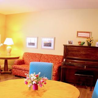 A cozy living room with a vibrant sofa, upright piano, and a table adorned with flowers.