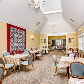 A cozy dining room with multiple tables and chairs, featuring a decorative quilt on the wall and a chandelier hanging from the ceiling.