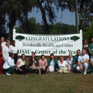 A group of people celebrating the Brooksville Healthcare Center being named HSM's Center of the Year, outdoors with a congratulatory banner.