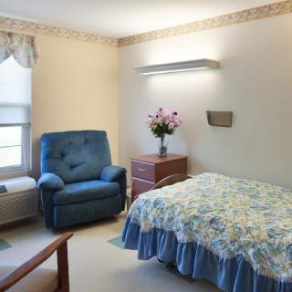 A cozy bedroom with a blue upholstered chair, floral bedding, and a vase of flowers on the nightstand.
