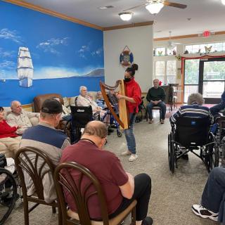 A harpist performs for residents in a nursing home, creating a lively atmosphere in a room with a nautical-themed wall mural.