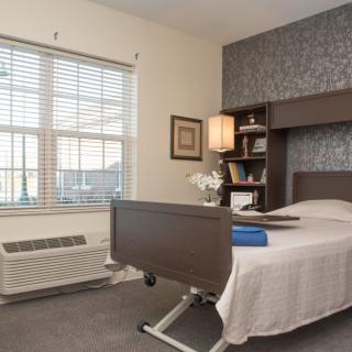 A hospital room with a single bed, bookshelf, and window letting in natural light.