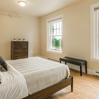 A minimalist bedroom with neutral colors, featuring a bed, dresser, and ample natural light from two windows.