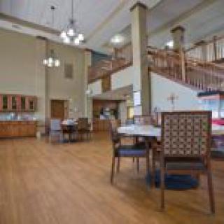 A spacious dining area with wooden flooring, featuring tables and chairs under high ceilings adorned with lighting fixtures.