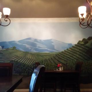 A cozy restaurant interior featuring a mural of rolling vineyards under chandeliers.