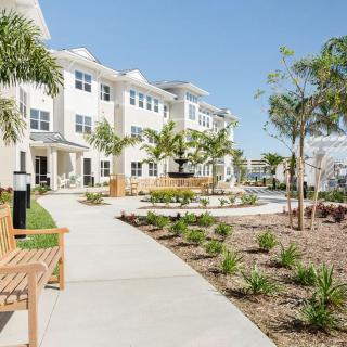 A modern apartment complex with a well-maintained pathway and landscaped garden featuring palm trees and benches.