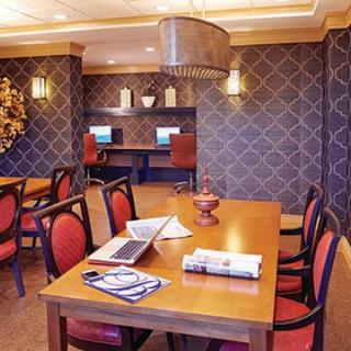A well-decorated dining room featuring a wooden table with chairs, a laptop, and books. In the background, there are computer workstations against a wall with patterned wallpaper and elegant lighting fixtures.
