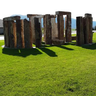 A view of a stone circle, reminiscent of ancient structures, surrounded by lush green grass under clear blue skies.