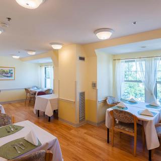 A brightly lit dining room with neatly arranged tables and chairs, featuring large windows that allow natural light to fill the space.