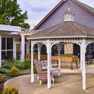 A peaceful garden setting with a white gazebo and comfortable seating, surrounded by landscaped greenery and a charming house.