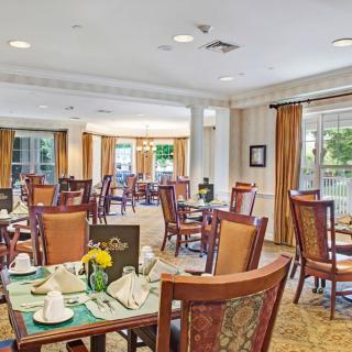 A spacious dining room with elegantly set tables and wooden chairs, featuring large windows and soft lighting.