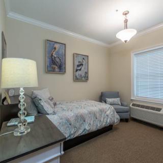 A cozy bedroom with a bed, chair, and wall art, softly lit by a lamp and natural light from the window.