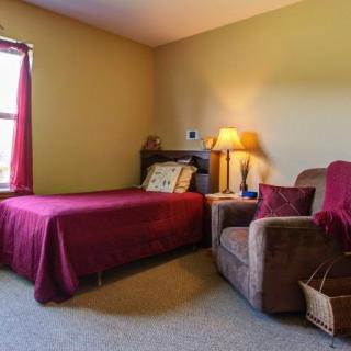 A cozy bedroom featuring a single bed with a maroon bedspread, an armchair beside a small lamp on a nightstand, and a window providing natural light.