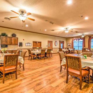 A spacious dining area with wooden floors, neatly arranged tables and chairs, ceiling fans, and warm lighting.