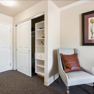 A cozy room featuring a white chair with a brown pillow, adjacent to an open closet and wall artwork.