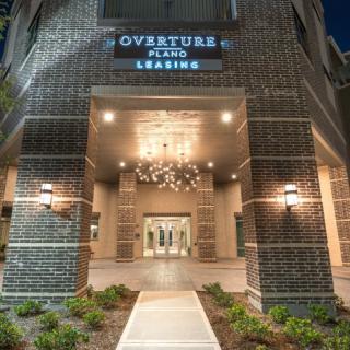 The illuminated entrance of Overture Plano at night, featuring modern architecture and a prominent chandelier.