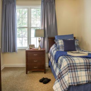 A cozy bedroom with a single bed featuring plaid bedding, a nightstand with a lamp, and curtains framing a window.