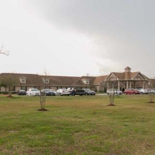 A large building with a parking lot in front, surrounded by a lawn and trees under a cloudy sky.