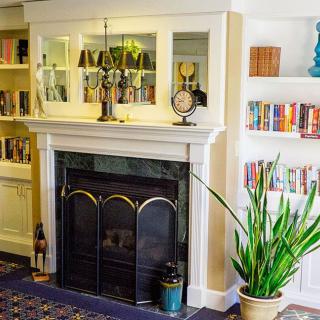 A cozy living room with a classic fireplace, surrounded by bookshelves and decorated with clocks and plants.