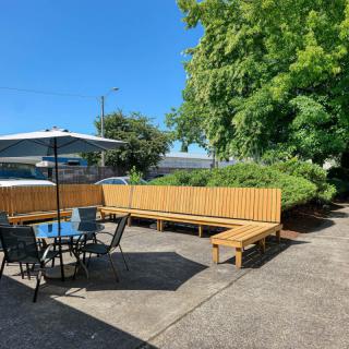 A sunny outdoor patio with a table, chairs, and benches under an umbrella surrounded by greenery.