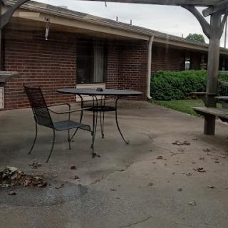 A quiet outdoor patio area with a table and chairs, surrounded by a brick building and greenery.