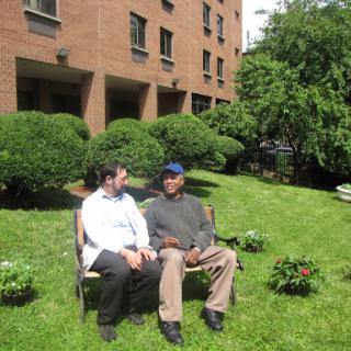 Two individuals engaged in a conversation on a bench in a grassy area surrounded by bushes.