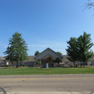 A building with a gabled roof, surrounded by trees, seen from across the road on a clear day.