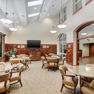 A spacious and well-lit community room featuring multiple tables and chairs, a large television, and skylights.