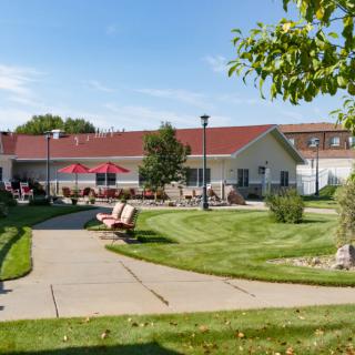 A scenic view of a building with a red roof, surrounded by a well-maintained lawn and pathway, featuring benches and trees.