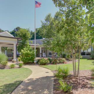 A building with a garden pathway, surrounded by trees and a flagpole on a sunny day.