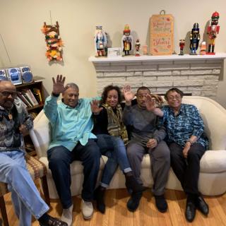 A group of people sitting on a couch in a cozy living room, smiling and waving at the camera.