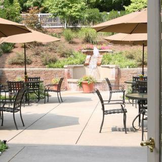 A serene outdoor patio area with tables, chairs, and umbrellas, featuring a central fountain surrounded by lush greenery.